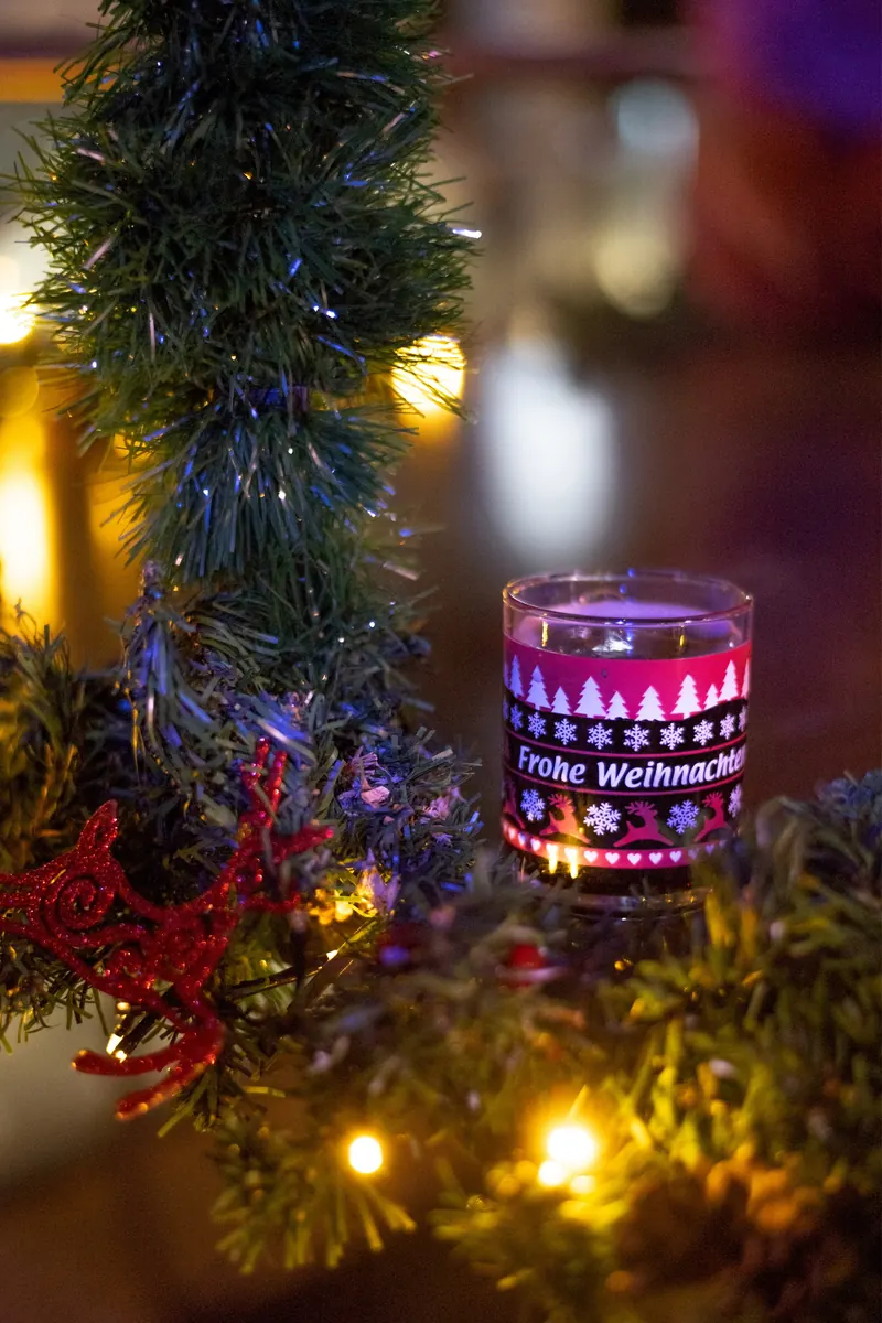 Verziertes Glas mit der Aufschrift „Frohe Weihnachten“ zwischen einer Tannengirlande mit Lichterketten